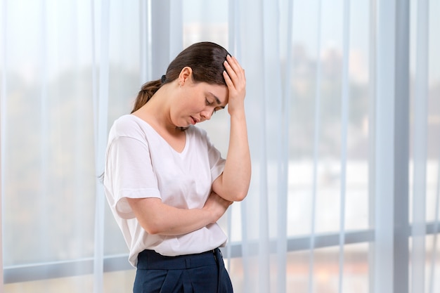 Portrait of crying woman holding her head with her hand. In the background of the window. Copy space. The concept of domestic violence.