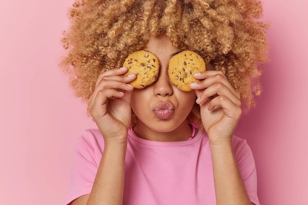portrait of culy haired young woman covers eyes with delicious cookies keeps lips folded dressed in casual t shirt isolated over pink background people confectionery and sweet food concept
