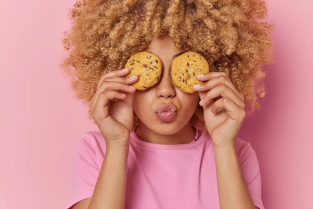 Portrait of culy haired young woman covers eyes with delicious cookies keeps lips folded dressed in casual t shirt isolated over pink background People confectionery and sweet food concept