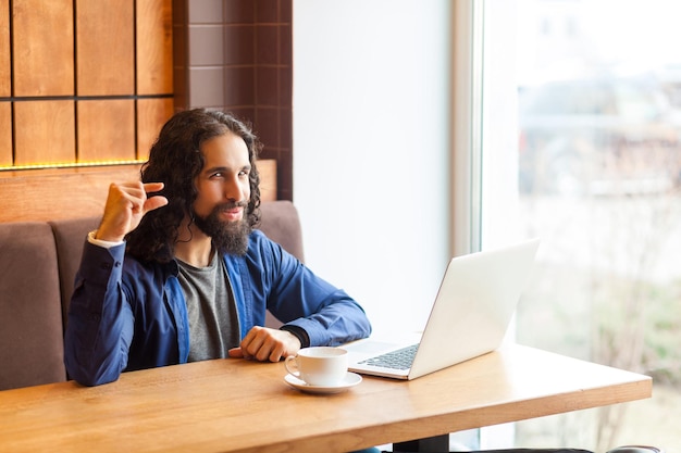 Portrait of cunning young adult man freelancer in casual style sitting in cafe with laptop, looking at camera, showing with finger to make less loud music. Indoor, lifestyle concept