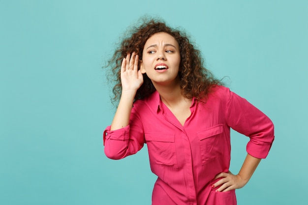 Portrait of curious african girl in casual clothes eavesdrop with hearing gesture isolated on blue turquoise wall background in studio. People sincere emotions, lifestyle concept. Mock up copy space.