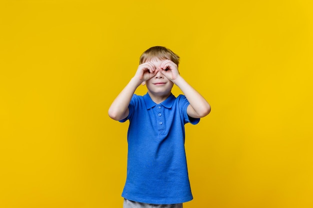 Portrait of a curious child looking through fingers in form of binoculars yellow isolated background