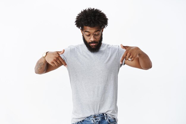 Portrait of curious handsome masucline african american male with beard and curly hair in glasses with tattoos, looking and pointing down with impressed and surprised expression on gray wall.