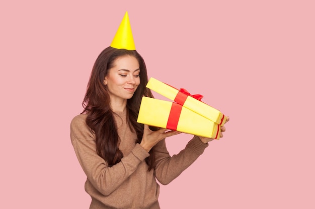 Portrait of curious happy woman with party cone on head looking inside box, unpacking gift and smiling pleased, satisfied with present, celebrating birthday, anniversary. indoor studio shot isolated