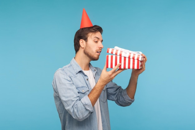 Ritratto di uomo curioso con regalo di apertura del cappello a cono di festa che dà una occhiata all'interno della scatola con un aspetto ficcanaso che disimballa presente in previsione di un'interessante sorpresa di compleanno girato in studio isolato su sfondo blu