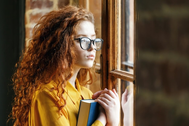 Portrait of curly ginger girl in the yellow shirt wearing glasses holding book and looking to the window at the loft placement