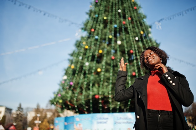 Portrait of a curly haired african woman wearing fashionable black coat and red turtleneck posing outdoor against main new year tree of city.