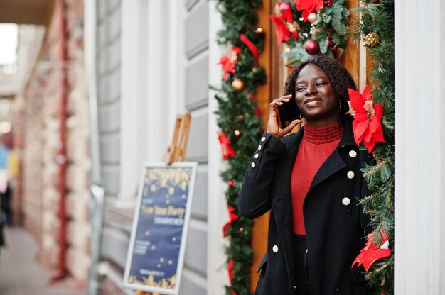 Portrait of a curly haired african woman wearing fashionable black coat and red turtleneck posing outdoor near door with christmas decoration, new year eve. Speak by phone.