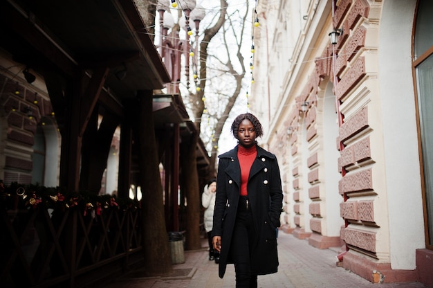 Portrait of a curly haired african woman wearing fashionable black coat and red turtleneck walking outdoor.