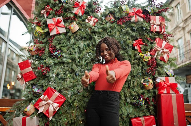 Portrait of a curly haired african woman wearing fashionable red turtleneck posing against christmas decorations, new year eve theme. Show thumbs up.