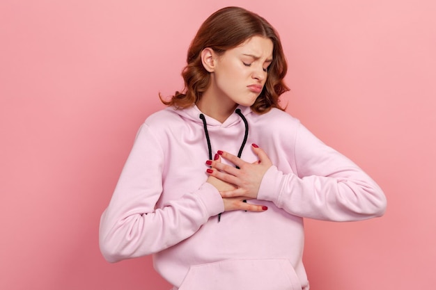 Portrait of curly haired teen girl in hoodie pressing on chest with painful expression, suffering sudden ache, heart attack, cardiac problems. Indoor studio shot isolated on pink background