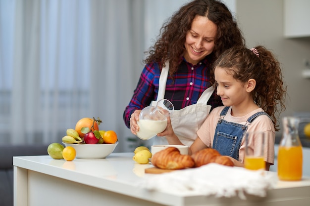 Il ritratto di una mamma riccia versa il latte della figlia carina in una ciotola con i cereali. è ora di colazione