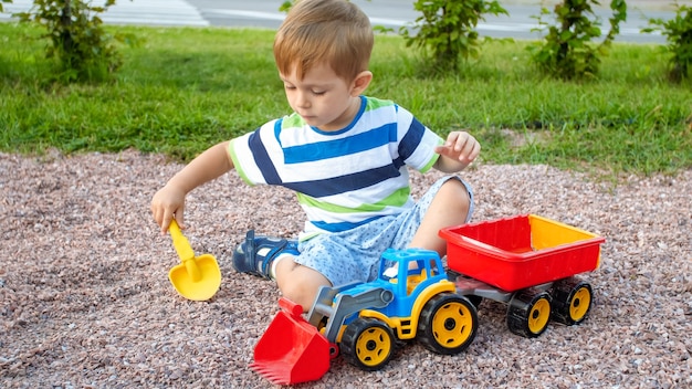 Portrait of cute 3 years old toddler boy sitting on the playground at park and playing with colorful plastic toy truck