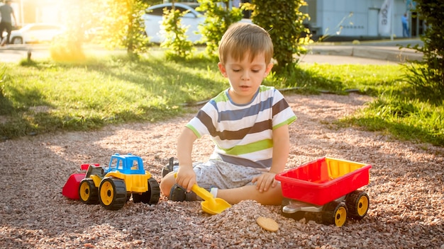 Portrait of cute 3 years old toddler boy sitting on the playground at park and playing with colorful plastic toy truck