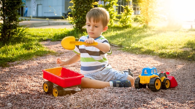 Portrait of cute 3 years old toddler boy sitting on the playground at park and playing with colorful plastic toy truck