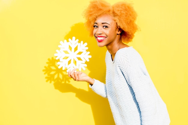 Portrait of a cute african woman in blue sweater holding an artificial snowflake on the yellow background