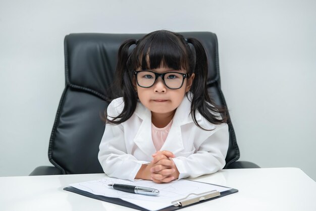 白い背景の上の机に取り組んでいる医者の制服を着たかわいいアジアの少女の肖像画