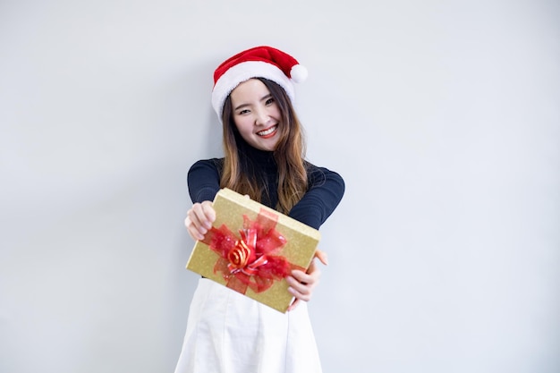 クリスマスの衣装を着たかわいいアジアの若い女性の肖像画と赤いサンタクロースの帽子をかぶって、白い背景とコピースペースに笑顔でギフトボックスを保持します。テーマクリスマスの十代の女性の幸せ。