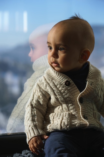 Portrait of a cute baby boy sitting on window sill with reflection in glass