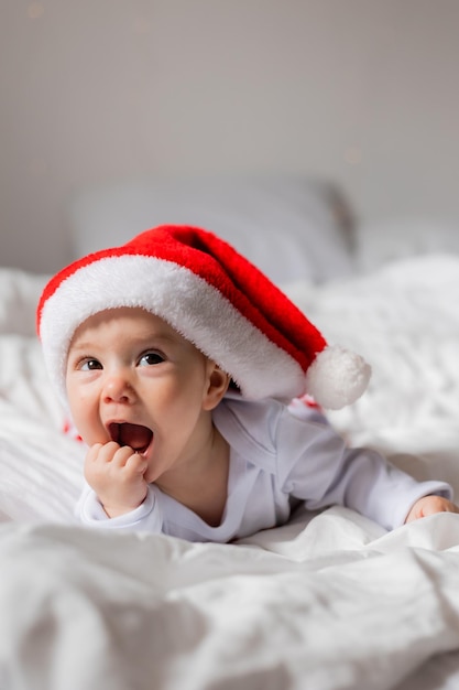 サンタの帽子をかぶった可愛い赤ちゃんの肖像画家でベッドで微笑んでいるクリスマス朝の高品質の写真