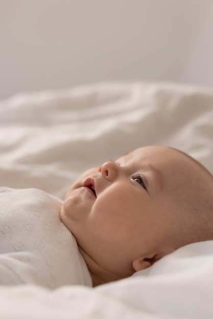 portrait of a cute baby in a white bodysuit on a bed at home with white linens. Newborn baby at home