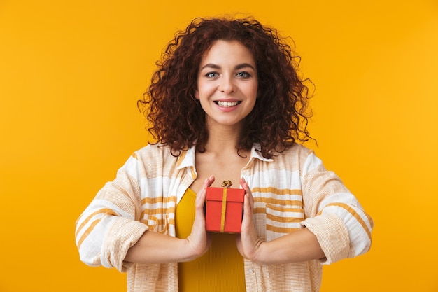 Portrait of a cute beautiful young curly woman posing isolated on yellow wall holding present gift box