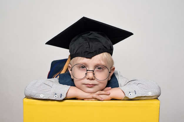 Portrait of a cute blond boy in big glasses, academic hat and a backpack. White background.