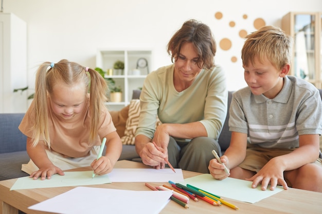 Portrait of cute blonde girl with down syndrome drawing with mother and brother together in home interior