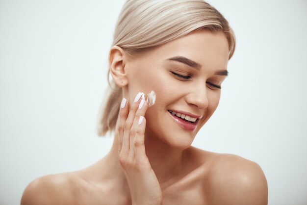 Portrait of cute blonde woman spreading cream on her face and smiling while standing against