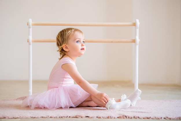 Portrait of cute blueeyed baby ballerina in pink sitting near the ballet barre putting on pointe shoes