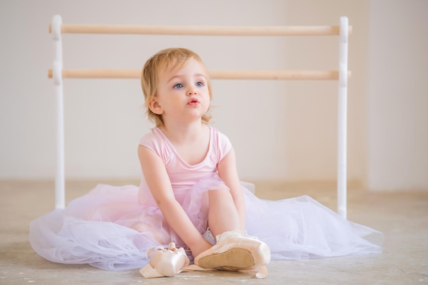 Portrait of cute blueeyed baby ballerina in pink sitting near the ballet barre putting on pointe shoes