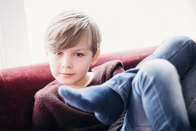 A portrait of a cute boy kid sitting on a sofa against the light window legs crossed