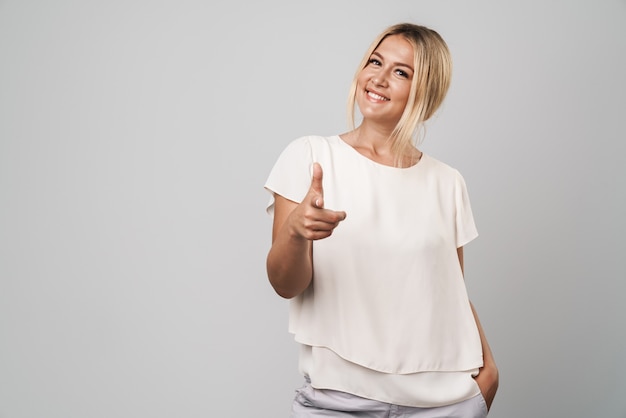 Portrait of a cute cheerful beautiful young amazing blonde woman posing isolated over grey wall dressed in basic white t-shirt pointing to you