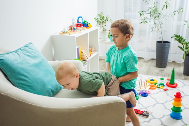 Portrait of cute cheerful brothers cooperate Elder brother holding younger one assisting him to climb the armchair Kids in their room with lots of toys Cooperation and support concept