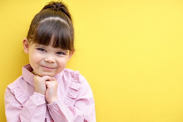 Portrait of cute child girl in pink blouse looking at camera posing isolated on yellow studio backgr...