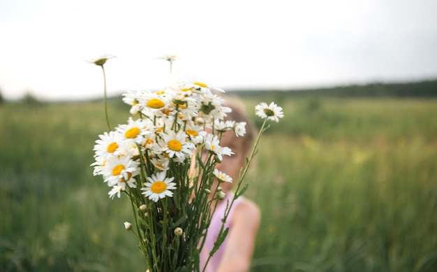 Portrait cute child girl with a bouquet of chamomile in summer\
on a green natural background happy child hidden face no face\
covered with flowers copy space authenticity rural life\
ecofriendly