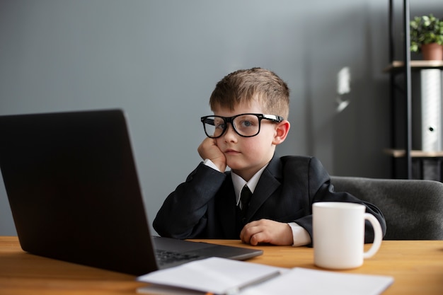 Photo portrait of cute child with suit sitting at the desk with laptop and mug