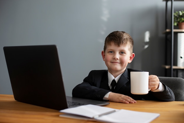 Portrait of cute child with suit sitting at the desk with laptop and mug