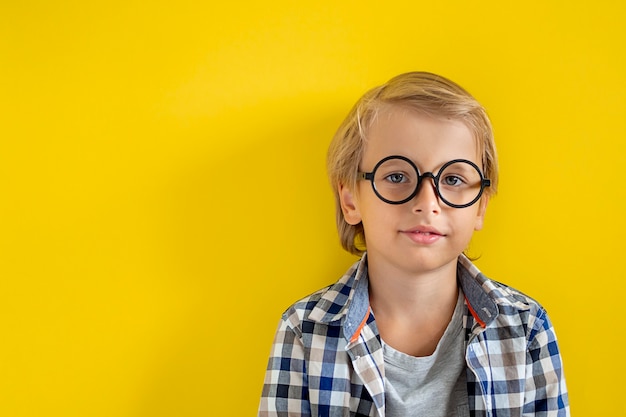 Portrait of cute and clever blonde Caucasian boy in a checked shirt on yellow background. 1 September day. Education and back to school concept. Child pupil ready to learn and study.