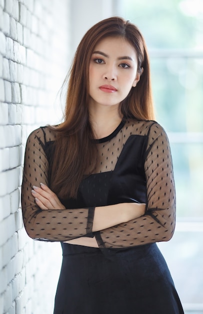 Portrait of cute content charming Asian female, Lao ethnicity, in stylish black dress standing  with crossed arms in room and looking at camera with self confidence.