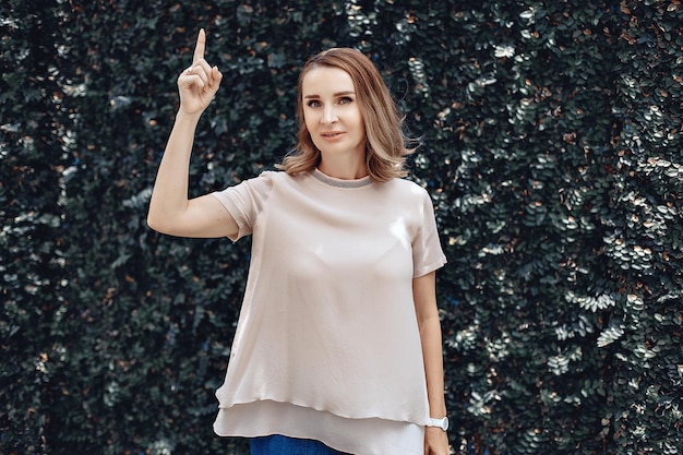 Portrait of cute decent smiling woman posing in pink t-shirt and showing index finger gesture against green leaves background