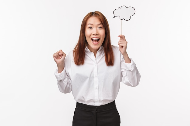 Portrait of cute and excited asian woman have great idea, being inspired, holding comment cloud on stick with beaming smile, sharing her thought, white wall