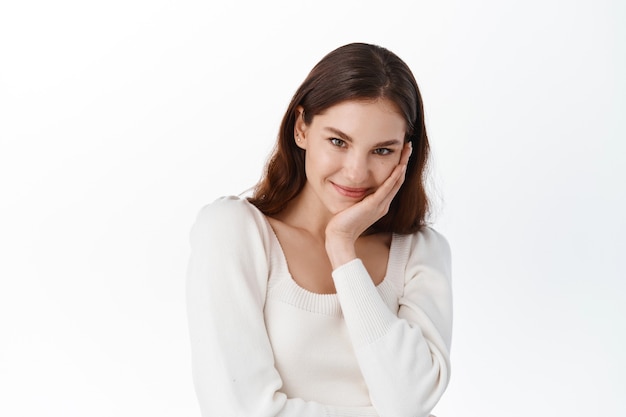 Portrait of cute female model in light blouse, giggle and looking shy, blushing from compliment, smiling at front modest, standing against white wall