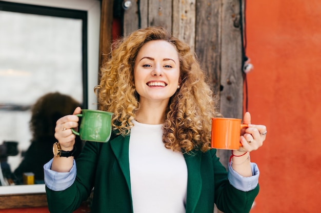 Portrait of cute female with pure skin having stylish hairstyle dressed in green jacket and white Tshirt holding two cups of tea going to give one to her boyfriend having pleased excited expression