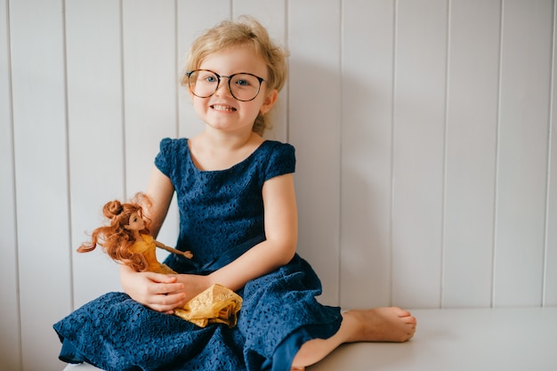 Portrait of cute and funny little girl in glasses holds her lovely barbie and posing over white wall