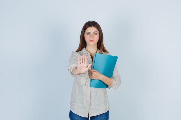 Portrait of cute Girl holding folder, showing stop gesture in shirt and looking serious