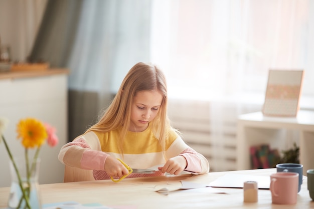 Portrait of cute girl making holiday card for Mothers day or Valentines day while sitting at table in cozy home interior, copy space