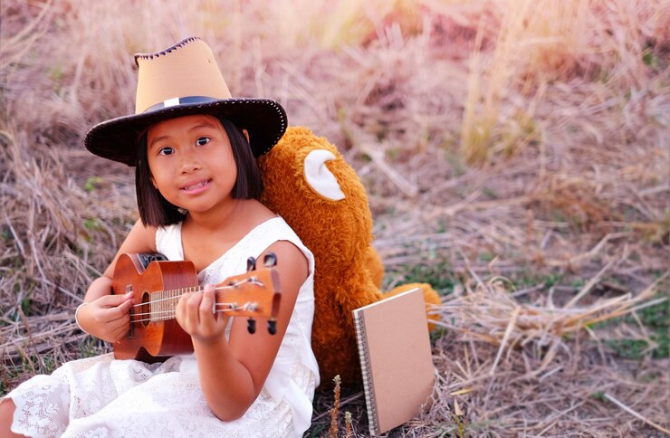 Photo portrait of cute girl playing guitar while sitting with book and teddy bear on field