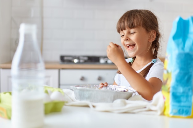 Portrait of cute girl taking baking, having happy facial expression, wearing white casual t shirt, posing indoor against kitchen set, being ready to taste delicious dessert.