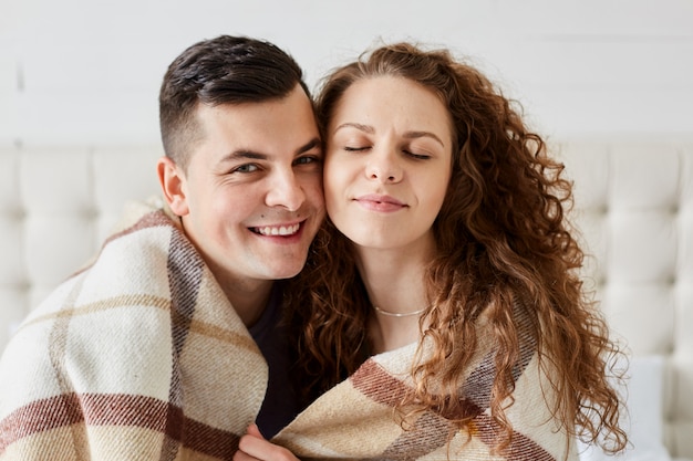 Portrait of cute happy couple sitting in bed embrace each other. Beautiful woman with long curly hair sits with closed eyes with her husband
